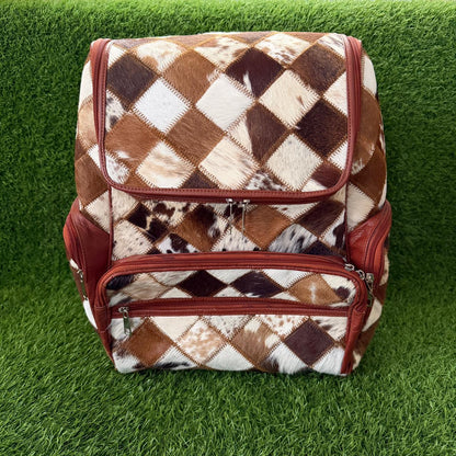 A backpack with a brown, beige, and white cowhide argyle pattern and reddish-brown leather trim, shown on a green grass background.