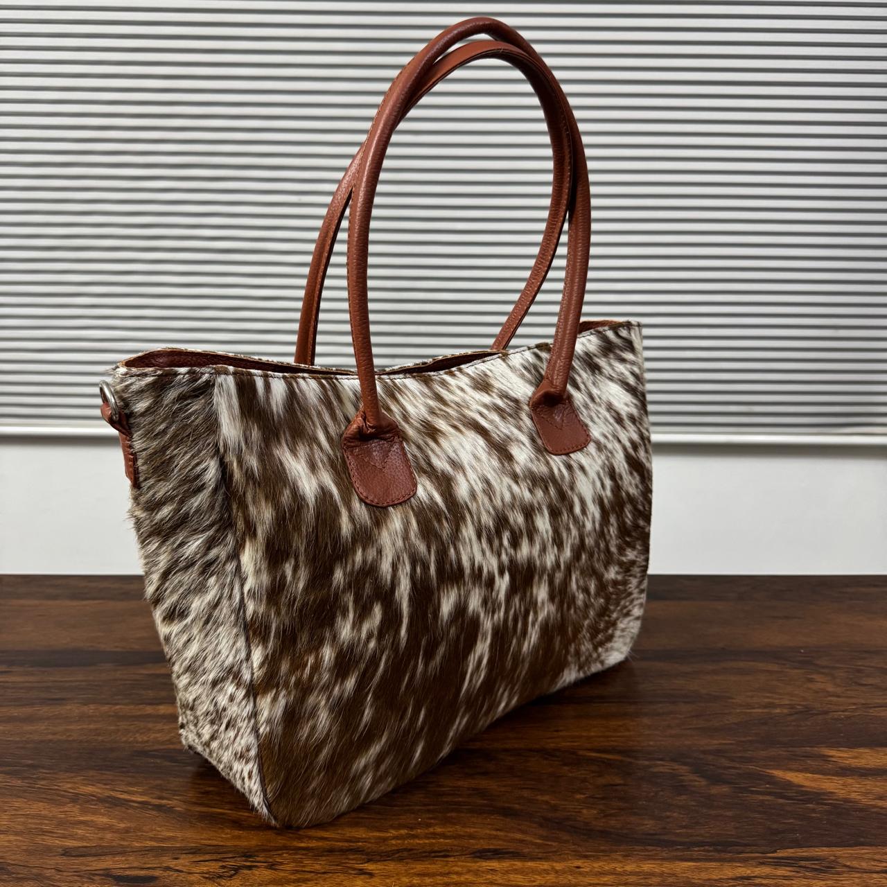 A cowhide handbag with brown leather handles, resting on a dark wooden table against a background of horizontal blinds.