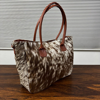 A cowhide handbag with brown leather handles, resting on a dark wooden table against a background of horizontal blinds.