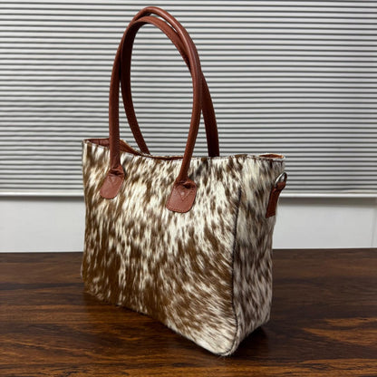 A cowhide tote bag with brown leather handles, sitting on a dark wooden table in front of a gray slatted window.