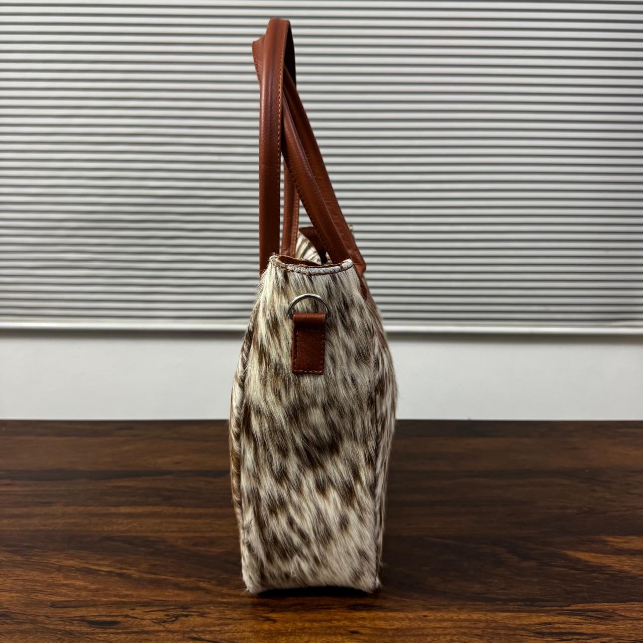 A structured tote bag with a brown and beige cowhide pattern body and brown leather handles, standing upright on a dark wooden surface against a gray slatted background.