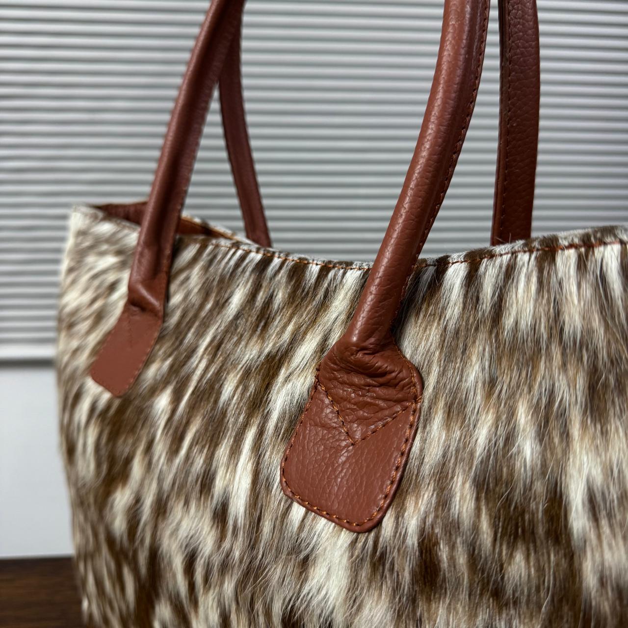 A close-up of a tote bag with a brown and beige animal-print faux fur body and smooth brown leather handles, set against a background of gray window blinds.