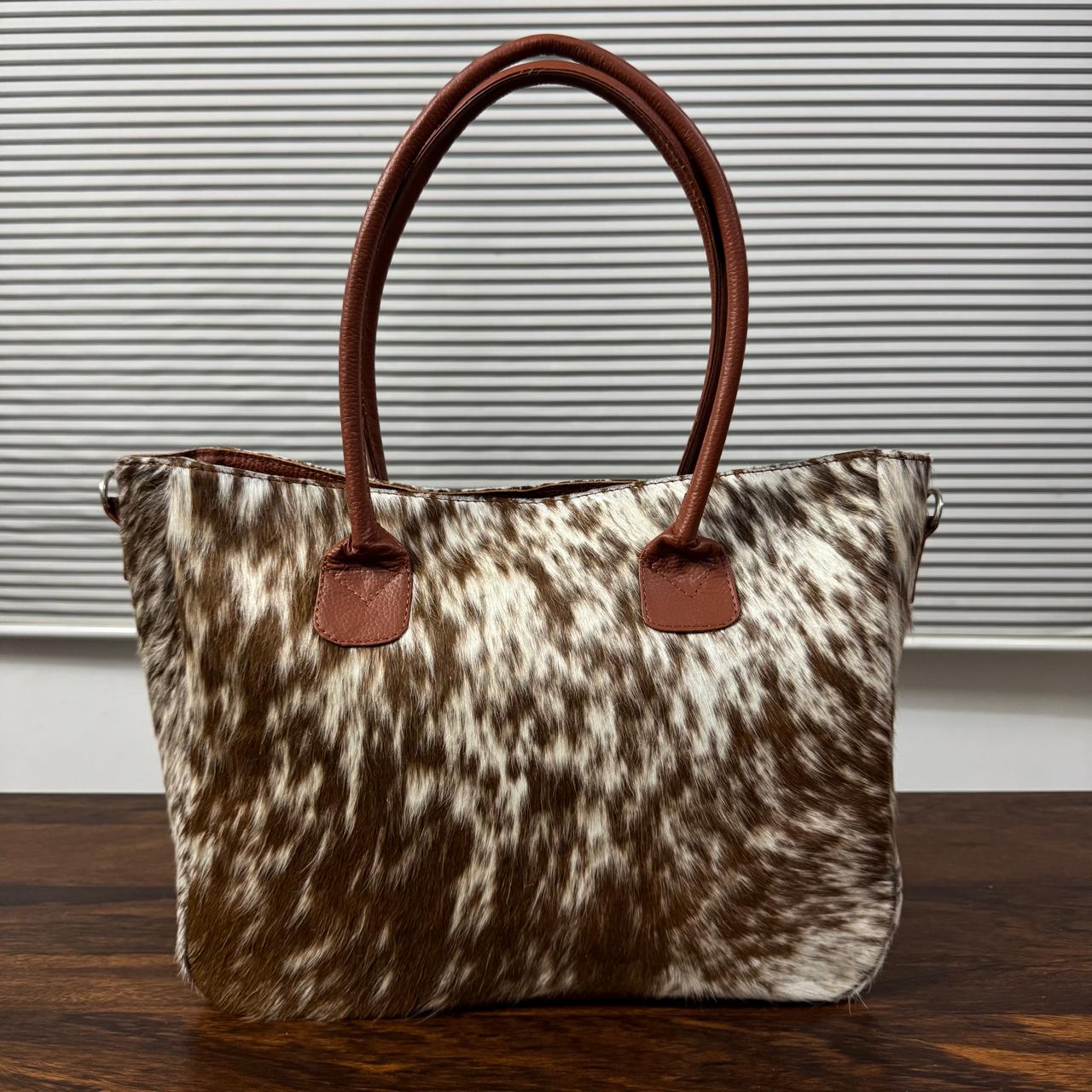 A cowhide handbag with brown leather handles and trim, displayed on a wooden table against a background of horizontal blinds.