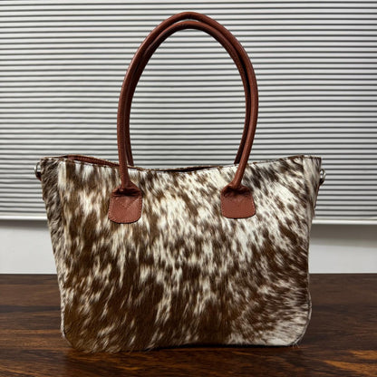 A cowhide handbag with brown leather handles and trim, displayed on a wooden table against a background of horizontal blinds.