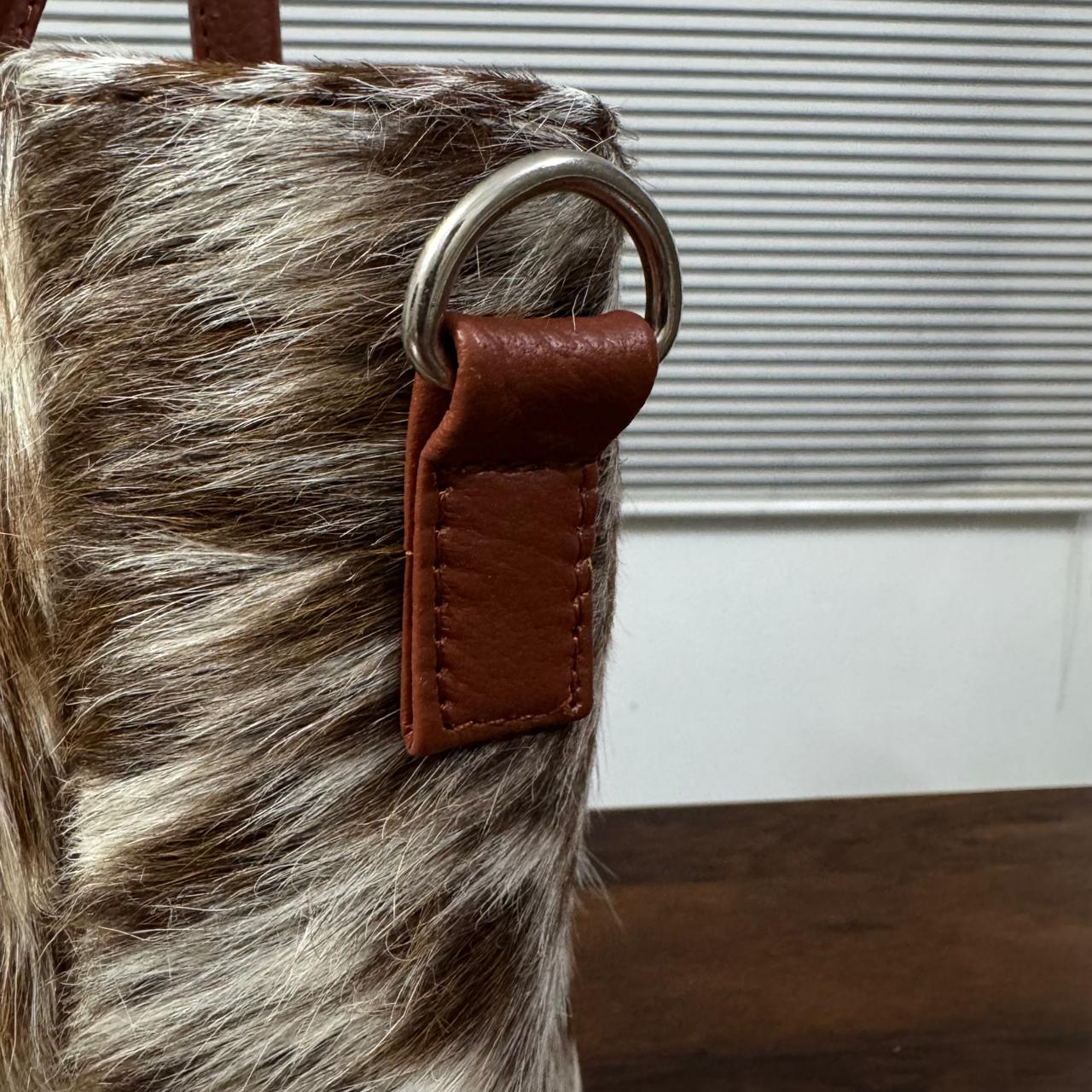Close-up of a brown leather strap attachment with a silver metal O-ring on a zebra-print bag, set against a background of window blinds and a wooden surface.