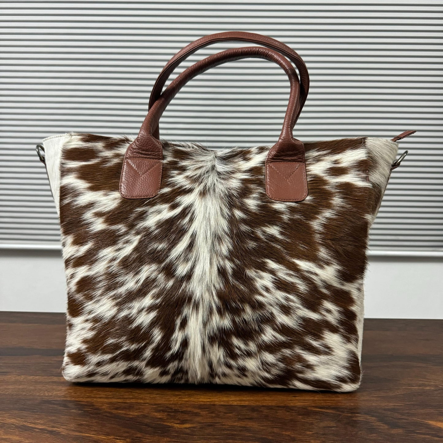 A cowhide tote bag with brown leather handles, resting on a dark wooden table in front of a gray slatted window background.