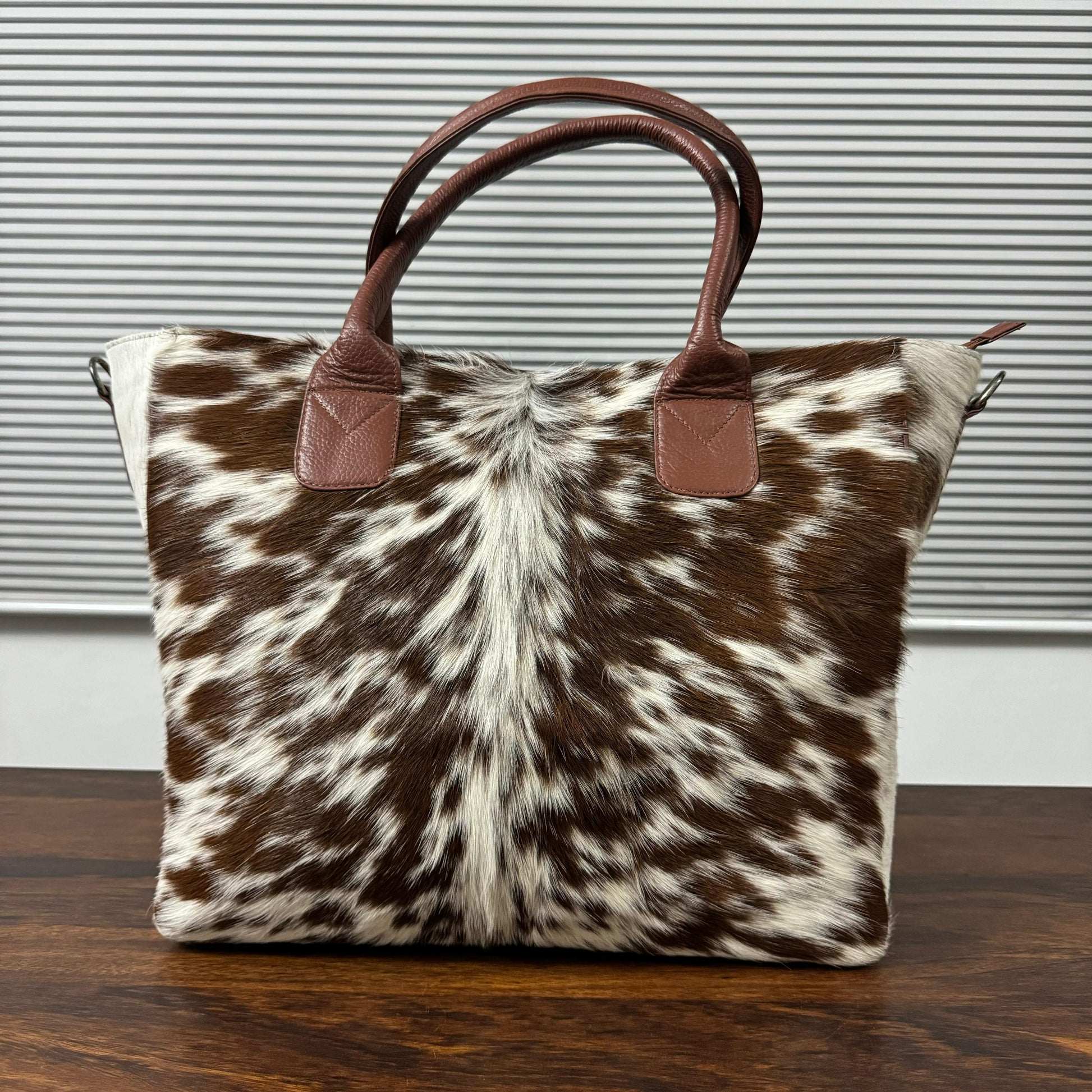 A cowhide tote bag with brown leather handles, resting on a dark wooden table in front of a gray slatted window background.