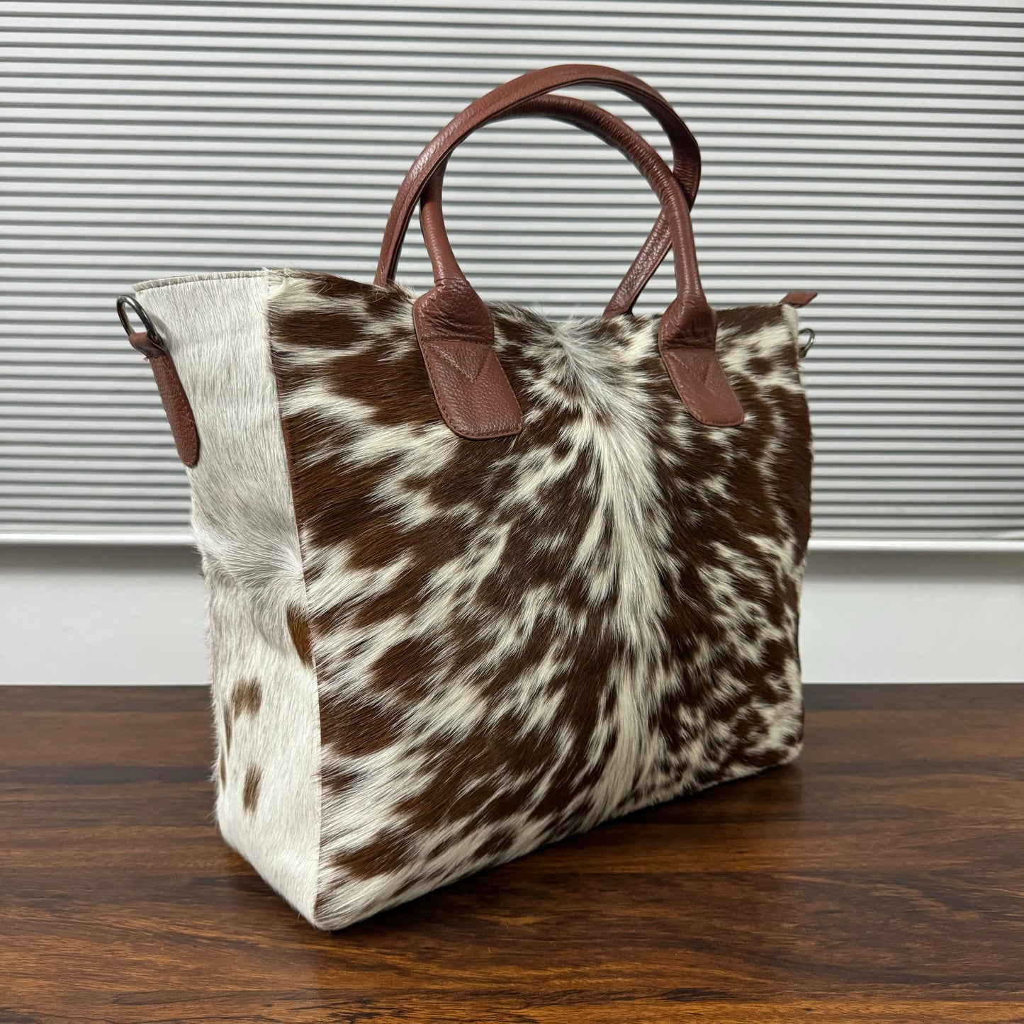 A cowhide tote bag with brown leather handles, resting on a dark wooden table in front of a gray slatted window.