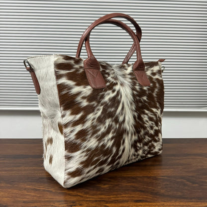 A cowhide tote bag with brown leather handles, resting on a dark wooden table in front of a gray slatted window.