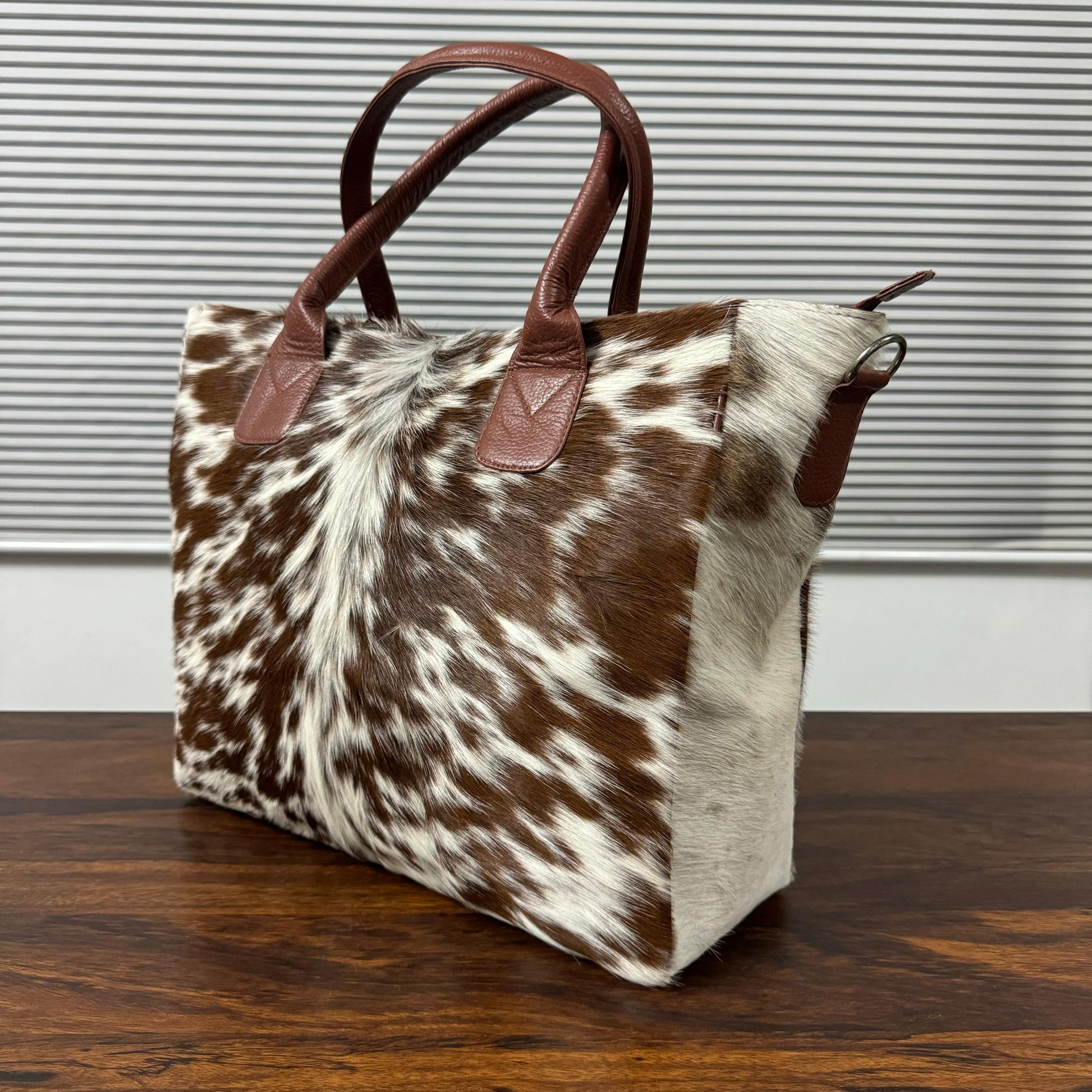 A cowhide tote bag with brown and white mottled pattern and dark brown leather handles, sitting on a dark wooden table in front of a gray slatted wall.