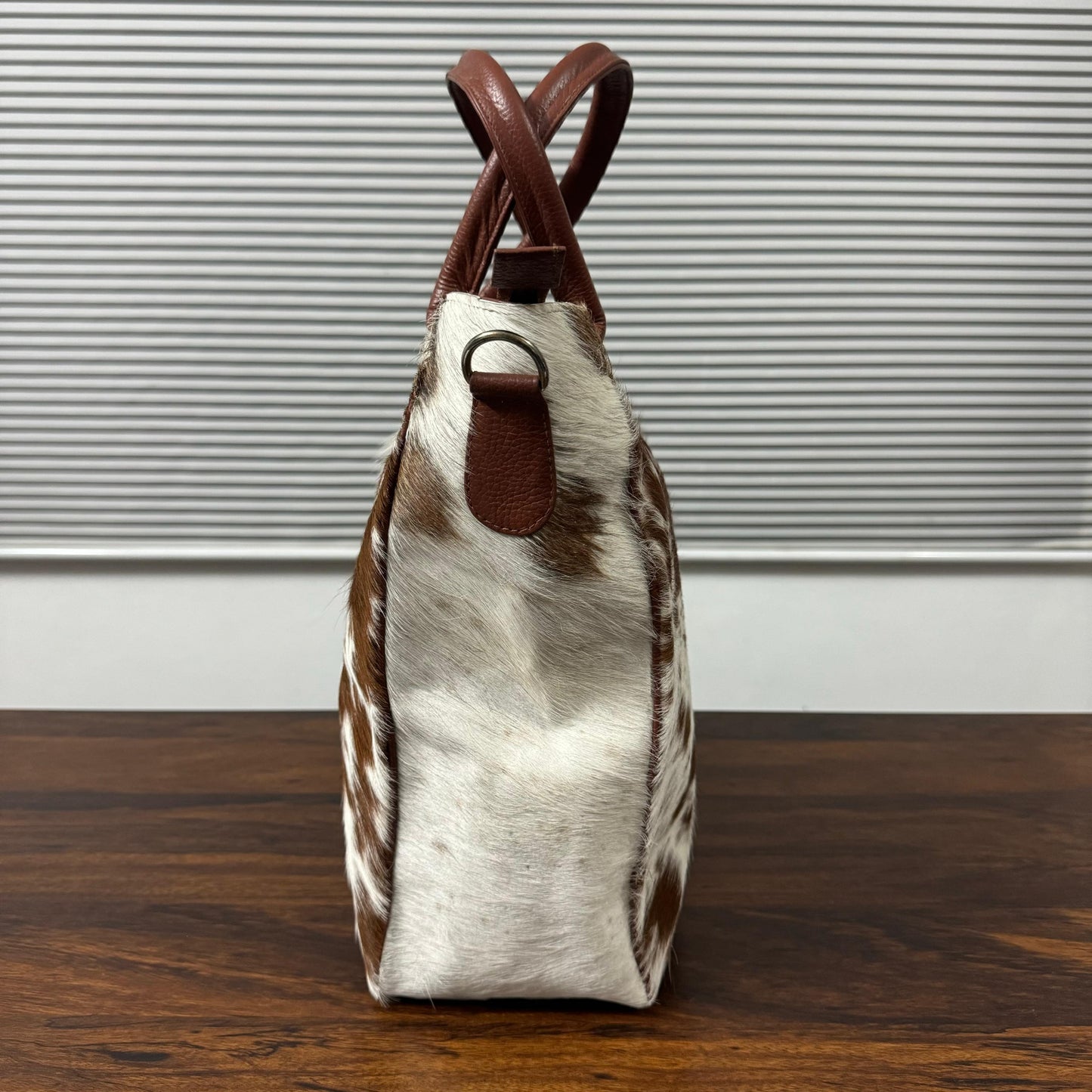 A structured handbag with a brown and white cowhide pattern body and brown leather handles, standing on a dark wooden table against a gray slatted wall.