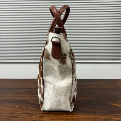 A structured handbag with a brown and white cowhide pattern body and brown leather handles, standing on a dark wooden table against a gray slatted wall.