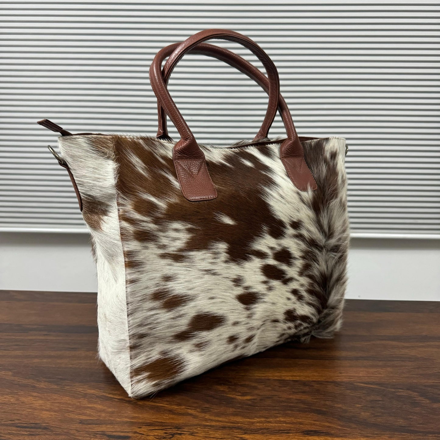 A cowhide tote bag with brown leather handles, sitting on a dark wooden table in front of a window with horizontal blinds.