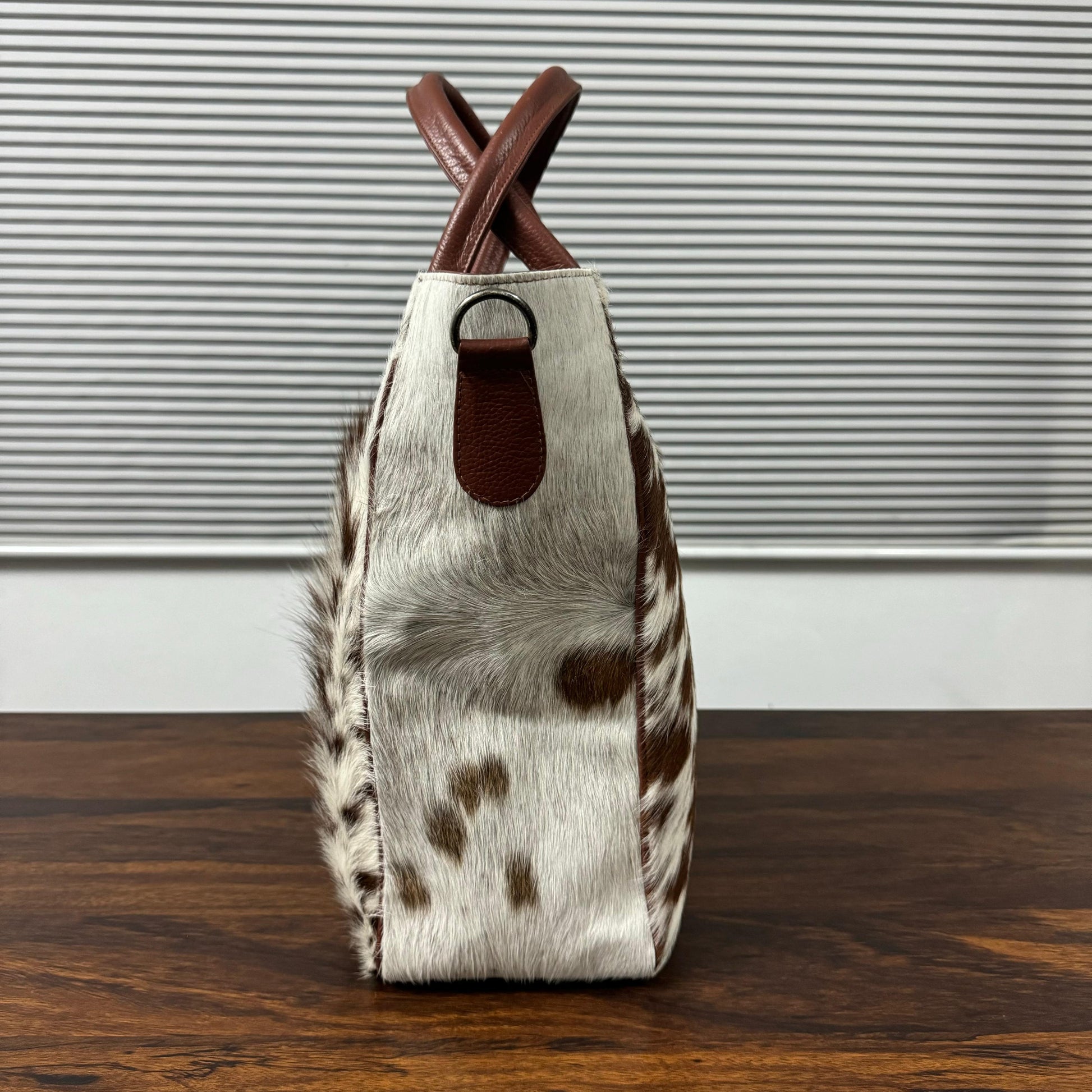 A structured handbag made of cowhide with brown and white spots, featuring brown leather handles and a matching brown leather accent tab, standing on a dark wooden surface in front of a gray slatted background.
