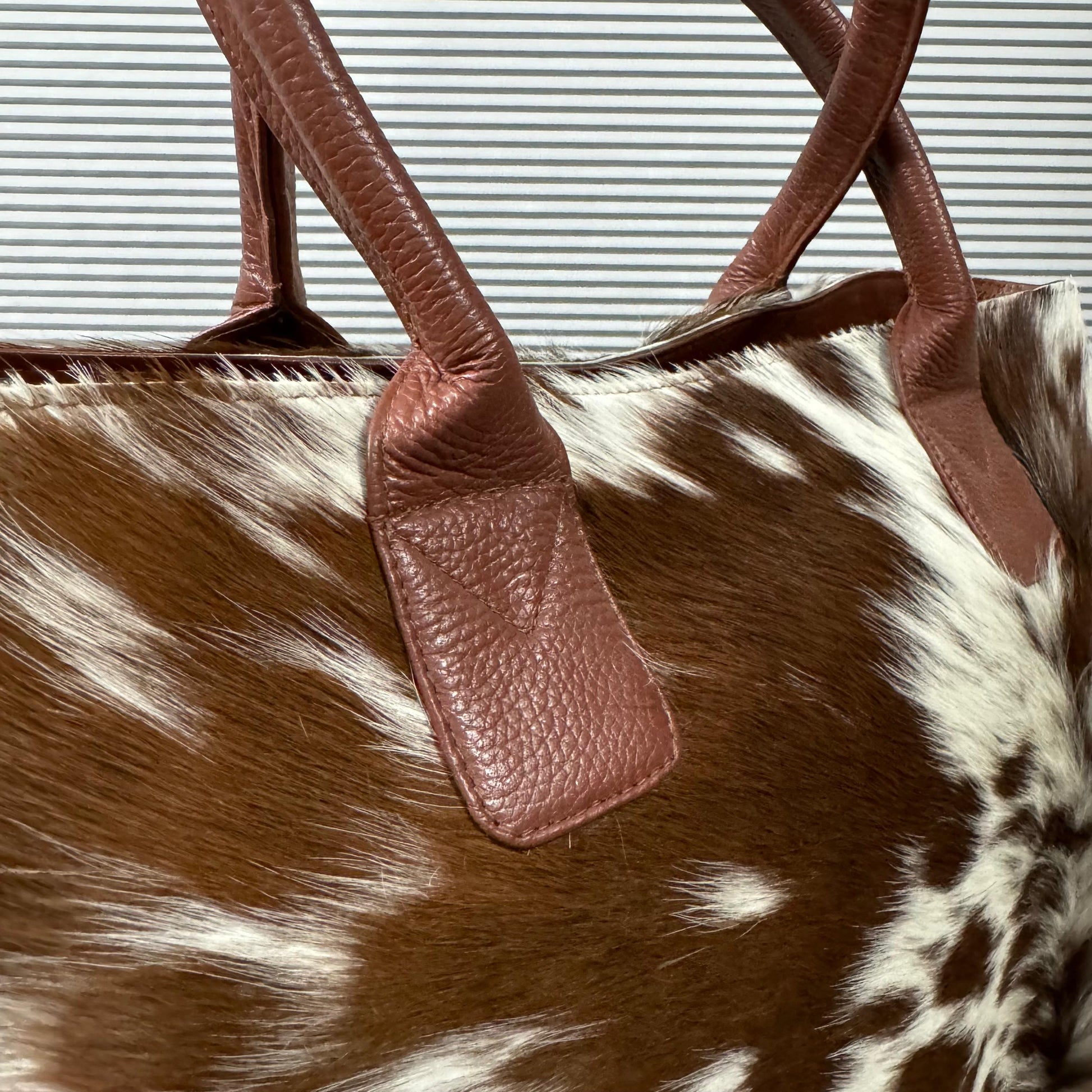 Close-up of a cowhide tote bag with brown and white spotted hide and brown leather handles, shown against a gray slatted background.
