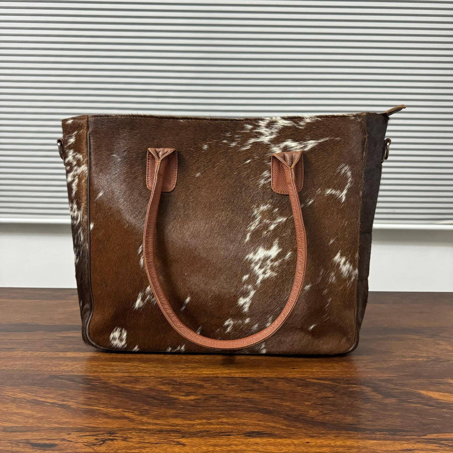 A brown and white cowhide tote bag with tan leather handles, standing on a wooden table in front of a window with horizontal blinds.