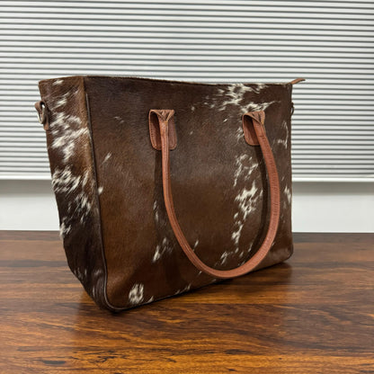 A brown and white cowhide tote bag with tan leather handles, resting on a wooden table in front of a window with horizontal blinds.