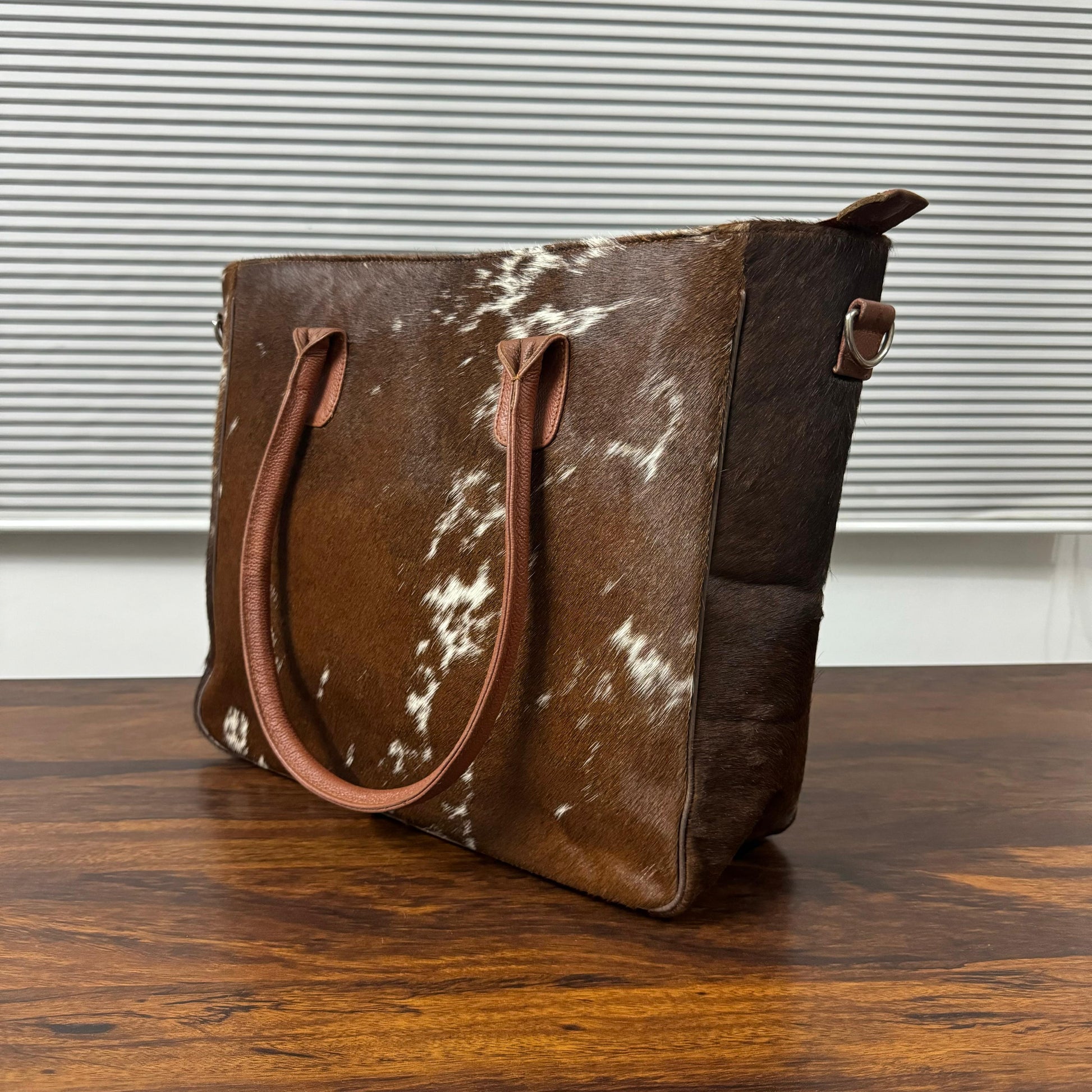 A brown and white cowhide tote bag with brown leather handles, standing on a wooden table in front of a window with horizontal blinds.