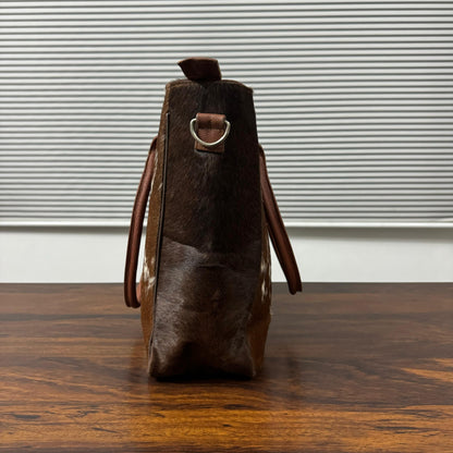 A brown cowhide shoulder bag with dark brown leather straps and a silver metal D-ring, standing upright on a wooden table in front of a window with horizontal blinds.