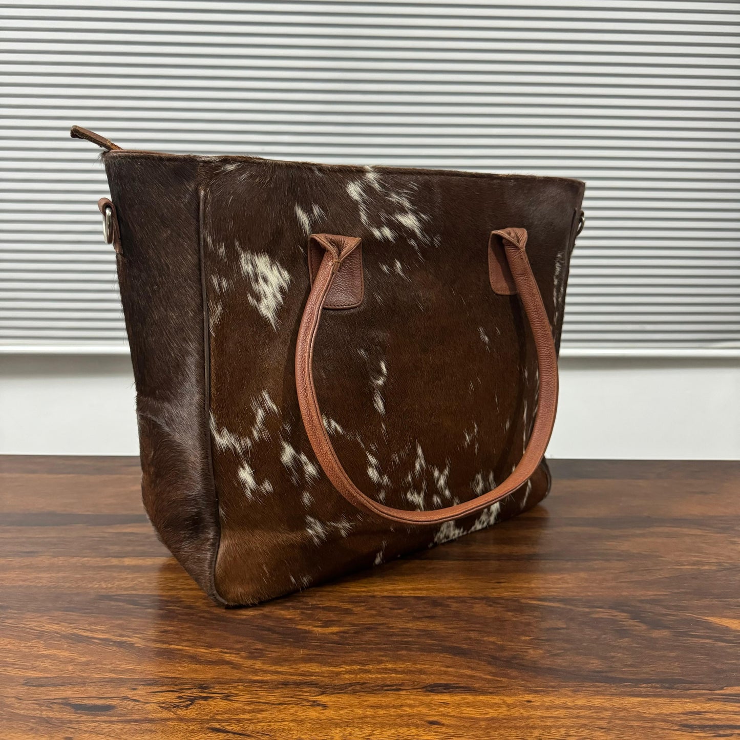 A brown and white cowhide tote bag with tan leather handles, sitting on a wooden table in front of a window with horizontal blinds.