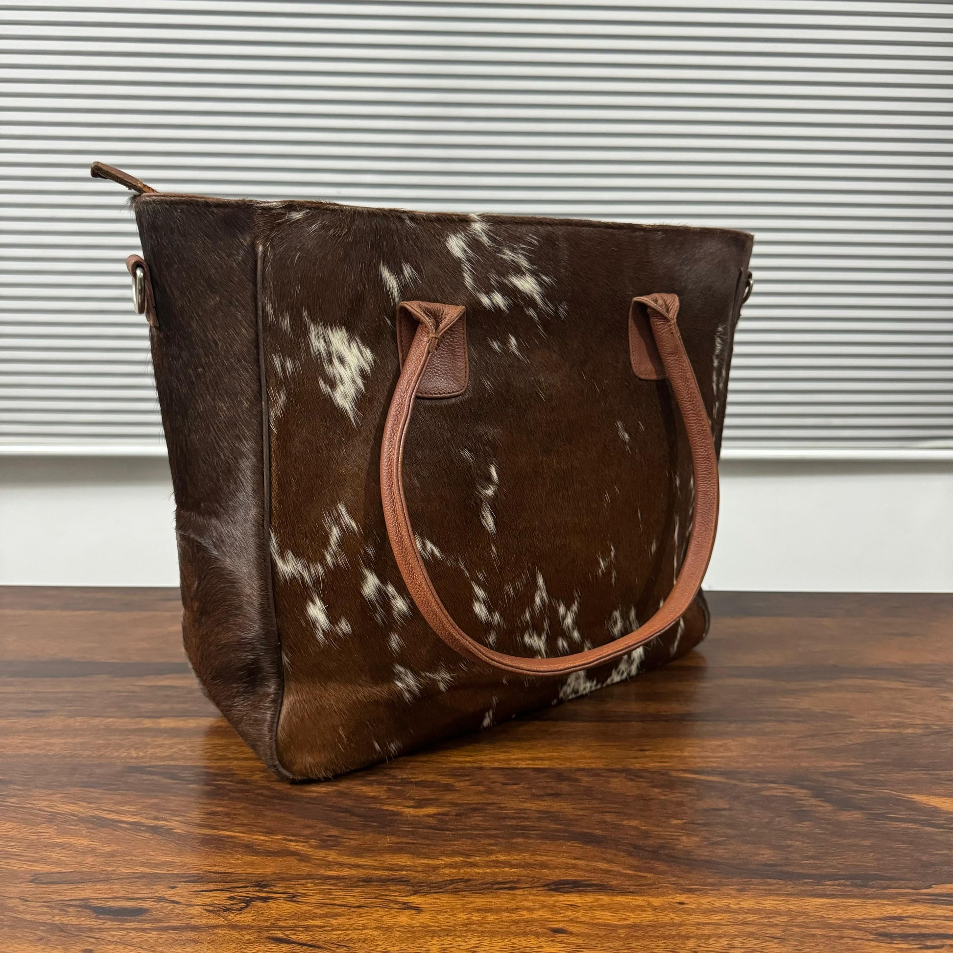 A brown and white cowhide tote bag with tan leather handles, sitting on a wooden table in front of a window with horizontal blinds.