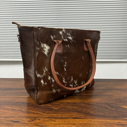 A brown and white cowhide tote bag with tan leather handles, sitting on a wooden table in front of a window with horizontal blinds.