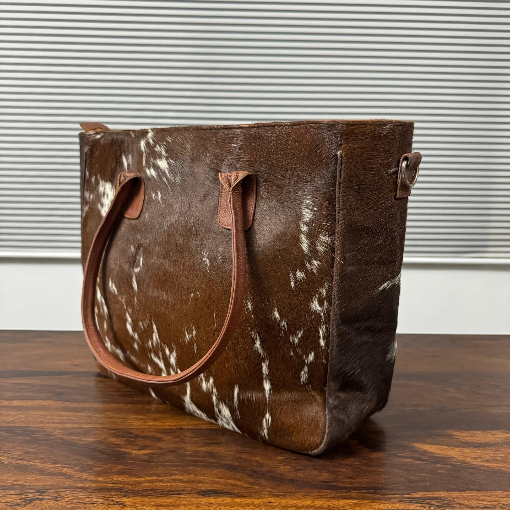 A brown and white cowhide tote bag with tan leather handles, sitting on a wooden table in front of a window with horizontal blinds.