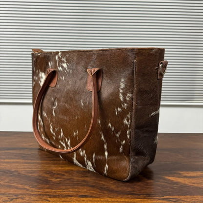A brown and white cowhide tote bag with tan leather handles, sitting on a wooden table in front of a window with horizontal blinds.