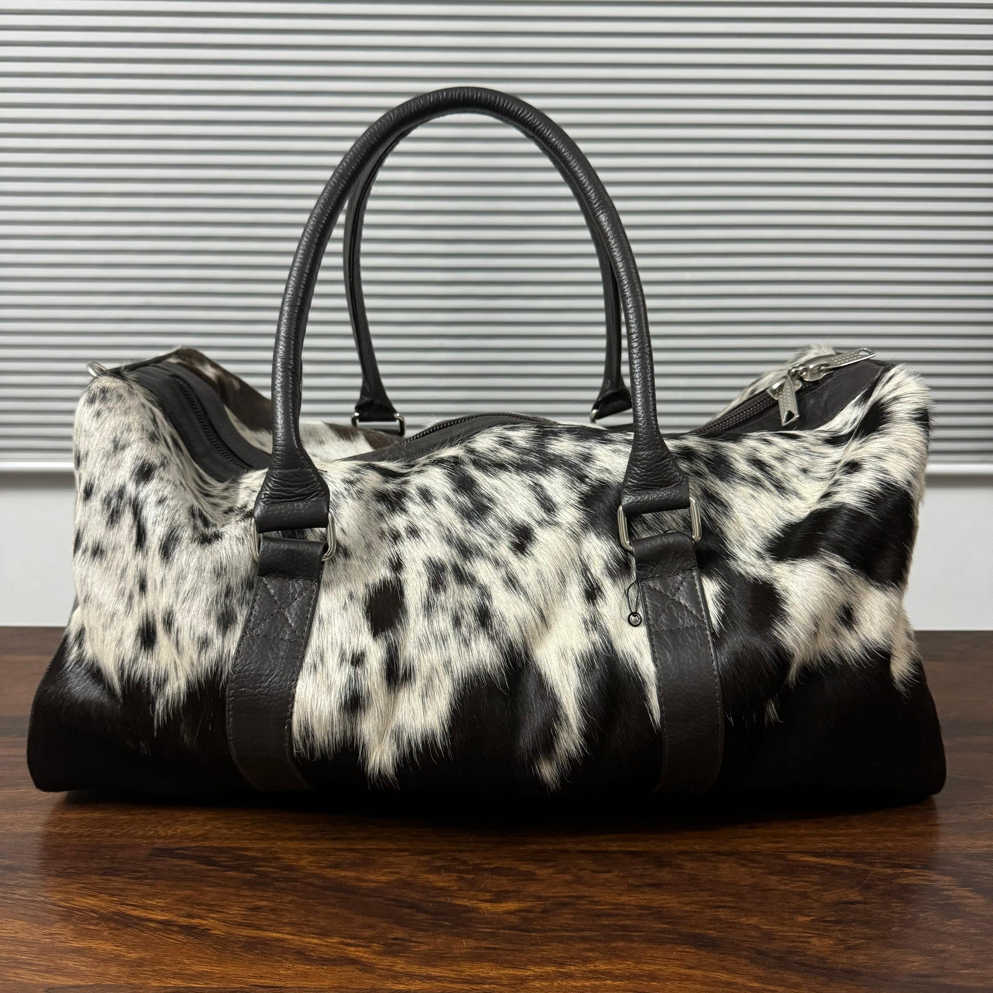 A black and white cowhide duffel bag with black leather handles and trim, resting on a dark wooden table against a gray slatted wall.