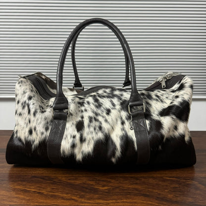 A black and white cowhide duffel bag with black leather handles and trim, resting on a dark wooden table against a gray slatted wall.