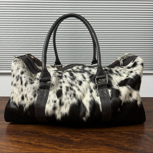A black and white cowhide duffel bag with black leather handles and trim, resting on a dark wooden table against a gray slatted wall.