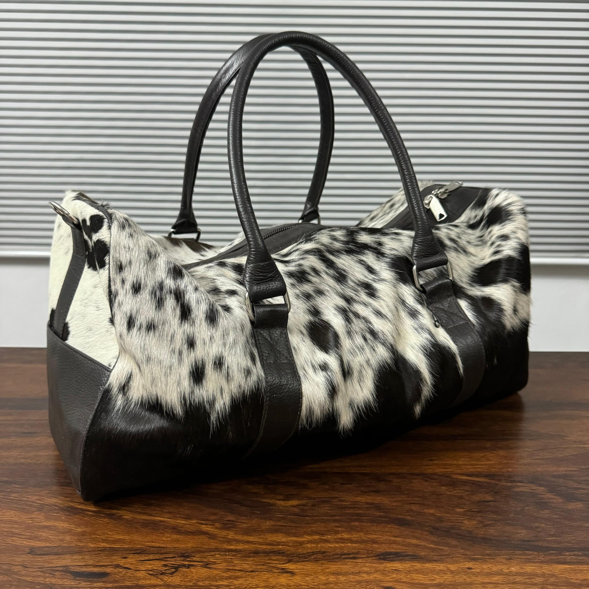 A duffle bag with a black and white cowhide pattern and black faux-leather trim, handles, and base, resting on a wooden table in front of a gray slatted wall.