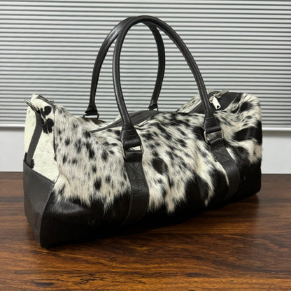 A duffle bag with a black and white cowhide pattern and black faux-leather trim, handles, and base, resting on a wooden table in front of a gray slatted wall.