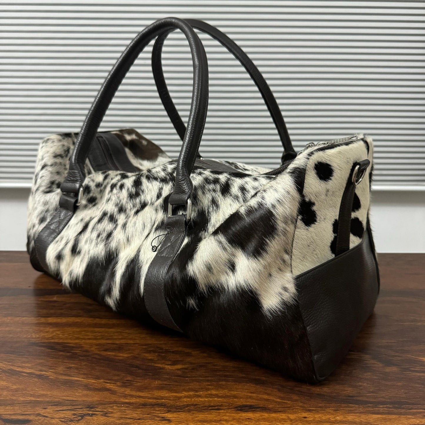 A duffle bag with a black and white cowhide pattern and black faux-leather trim, displayed on a wooden table in front of a window with horizontal blinds.
