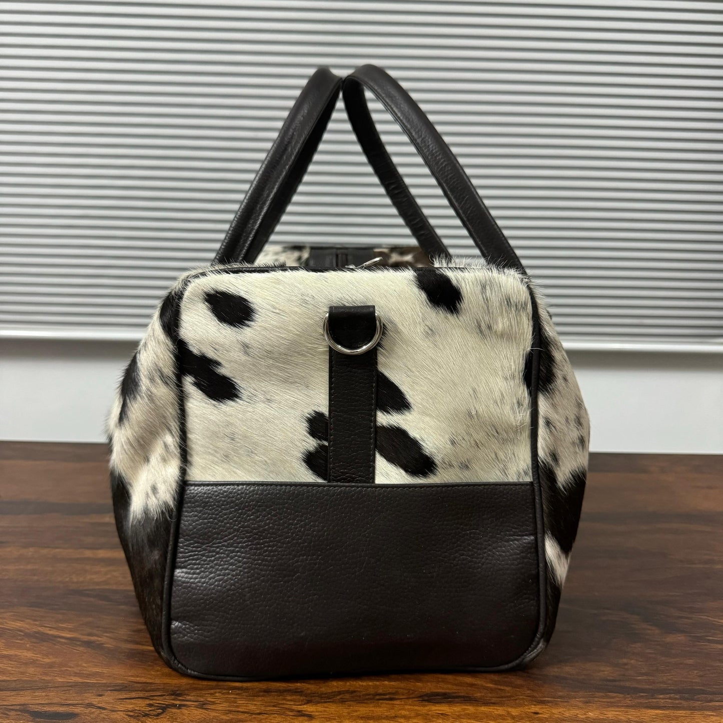 A cowhide and leather handbag with black spots on a white hide, black leather handles and base, and a black leather strap detail, sitting on a wooden table in front of a window with horizontal blinds.