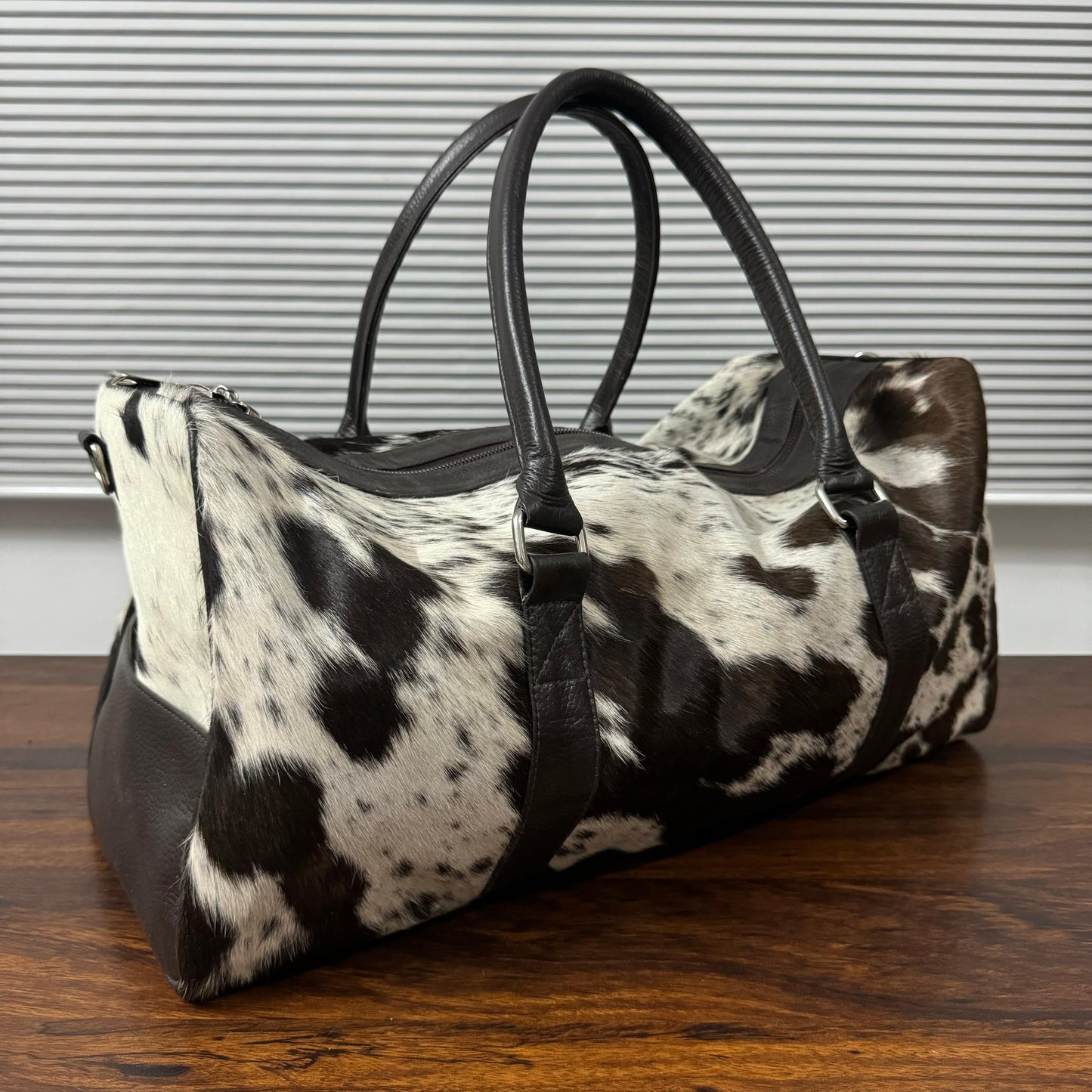 A cowhide duffel bag with black and white spotted pattern and black leather handles, resting on a dark wooden table in front of a window with horizontal blinds.