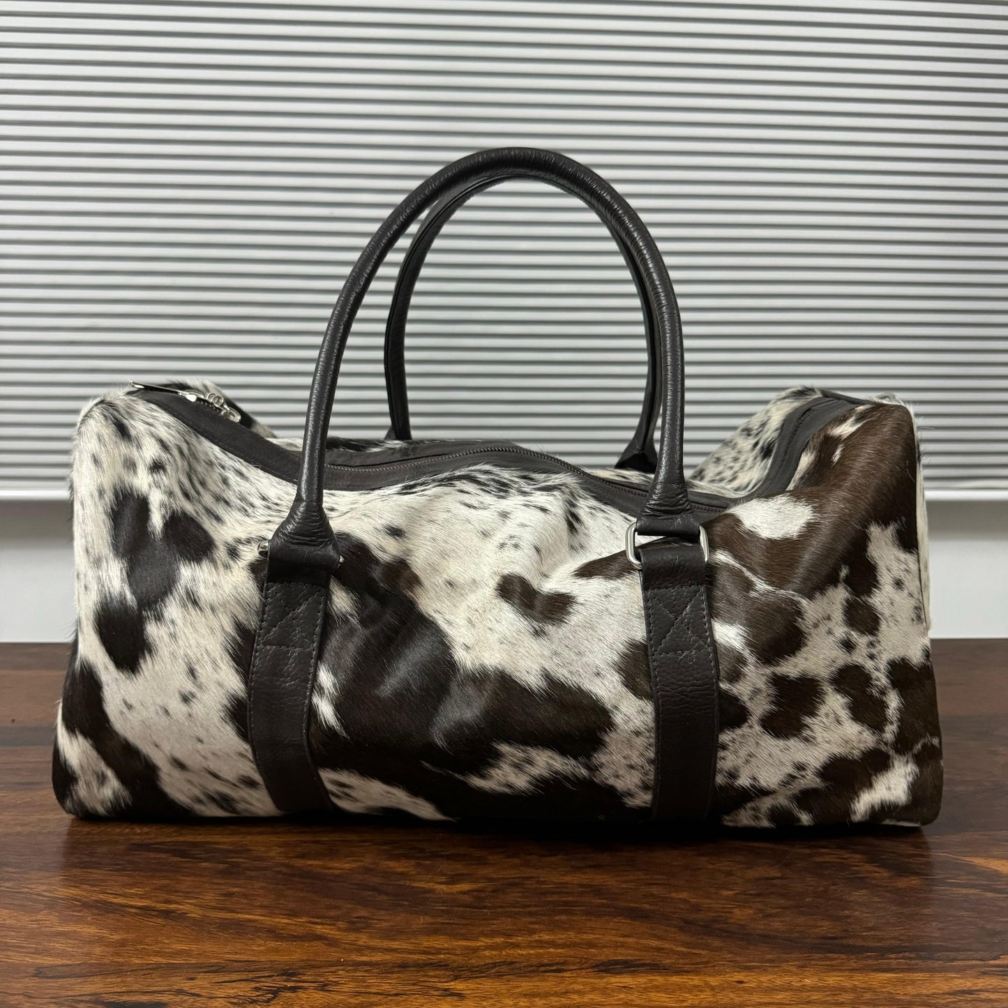 A duffle bag with a black and white cowhide pattern and black leather handles, sitting on a wooden table in front of a window with horizontal blinds.