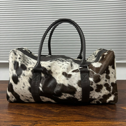 A duffle bag with a black and white cowhide pattern and black leather handles, sitting on a wooden table in front of a window with horizontal blinds.