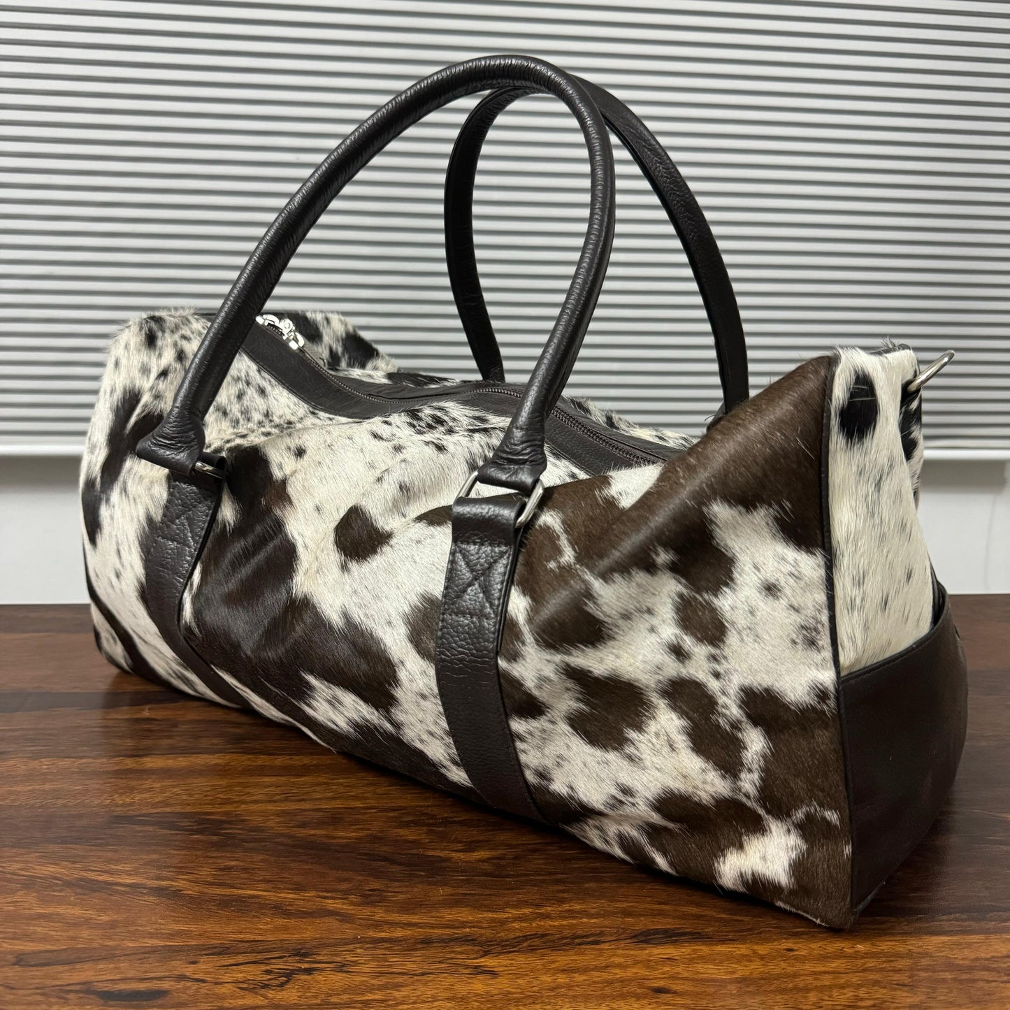 A cowhide duffel bag with black faux-leather handles and trim, resting on a wooden table in front of a window with horizontal blinds.