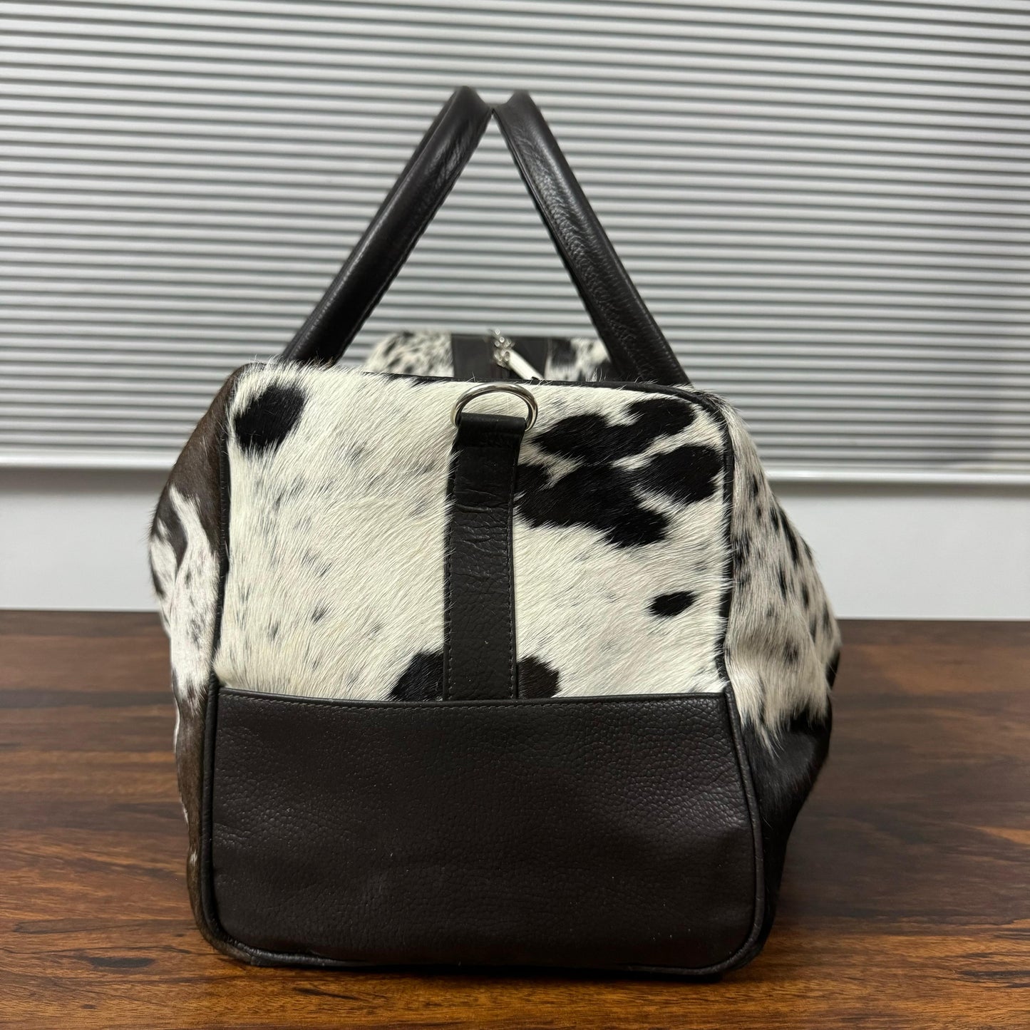 A cowhide and black leather duffel bag with black handles, resting on a wooden table in front of a window with horizontal blinds.