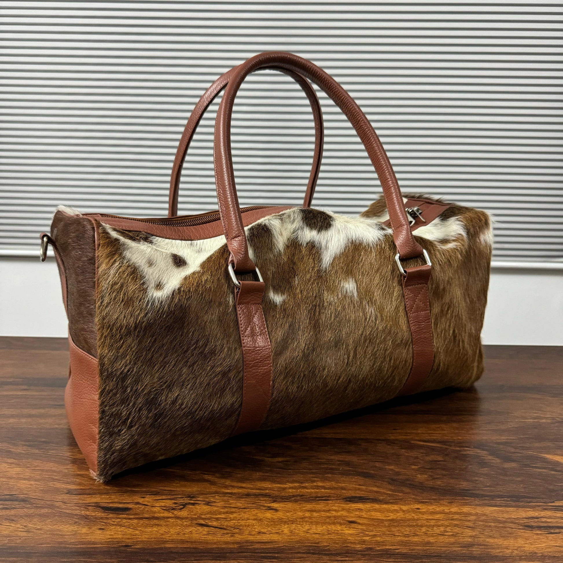 A cylindrical duffel bag with a brown and white cowhide body and tan leather handles and trim, resting on a dark wooden table in front of a gray slatted wall.