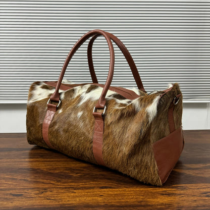 A duffle bag made of brown and white cowhide with brown leather handles and trim, resting on a wooden table in front of a window with horizontal blinds.
