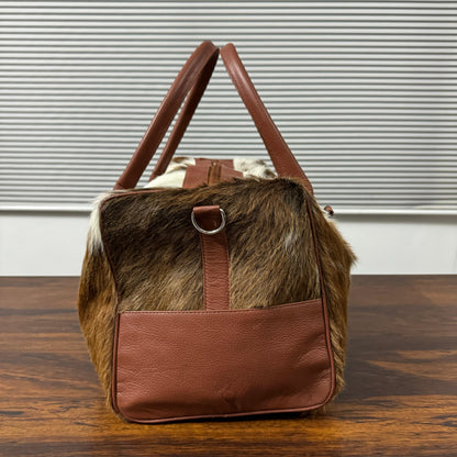 A brown and black cowhide duffel bag with brown leather handles and a matching front pocket, sitting on a wooden table in front of a window with horizontal blinds.