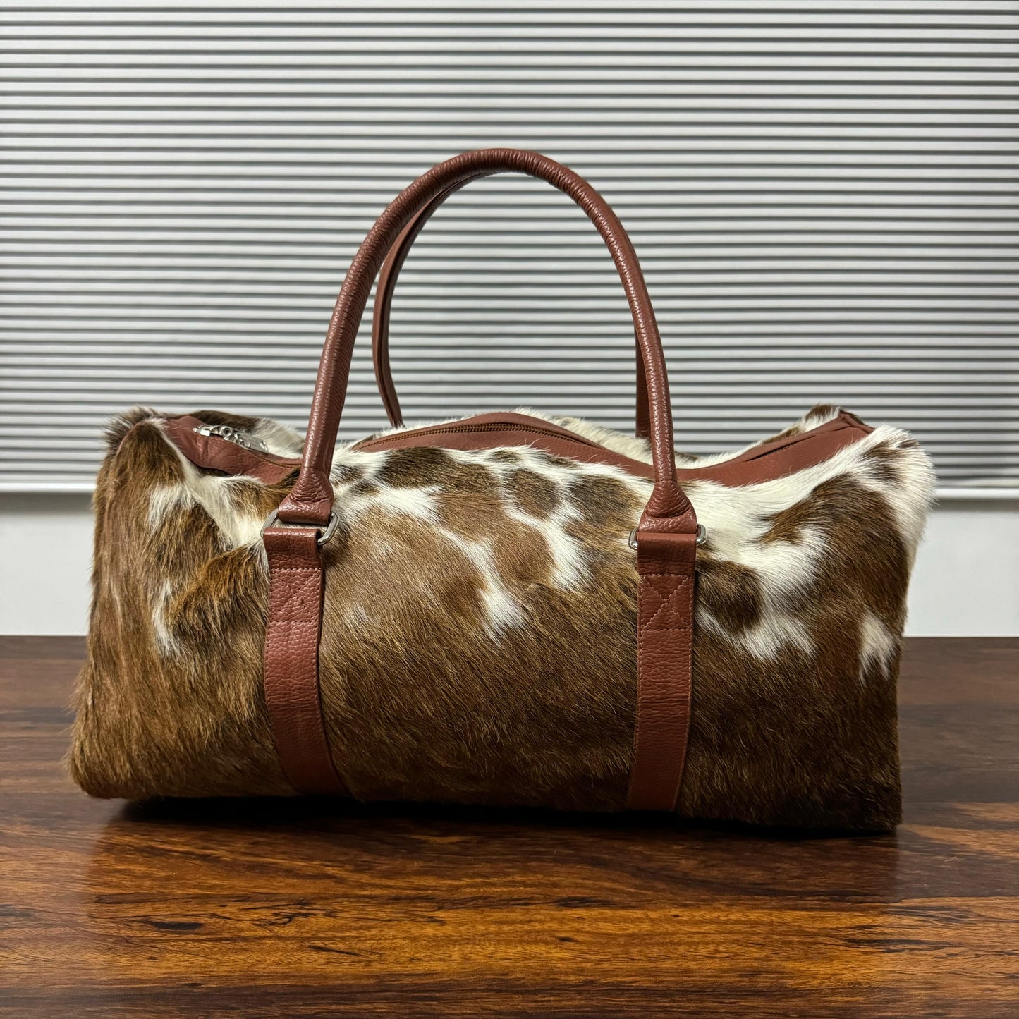 A cowhide duffel bag with brown leather handles and trim, resting on a wooden table in front of a window with horizontal blinds.