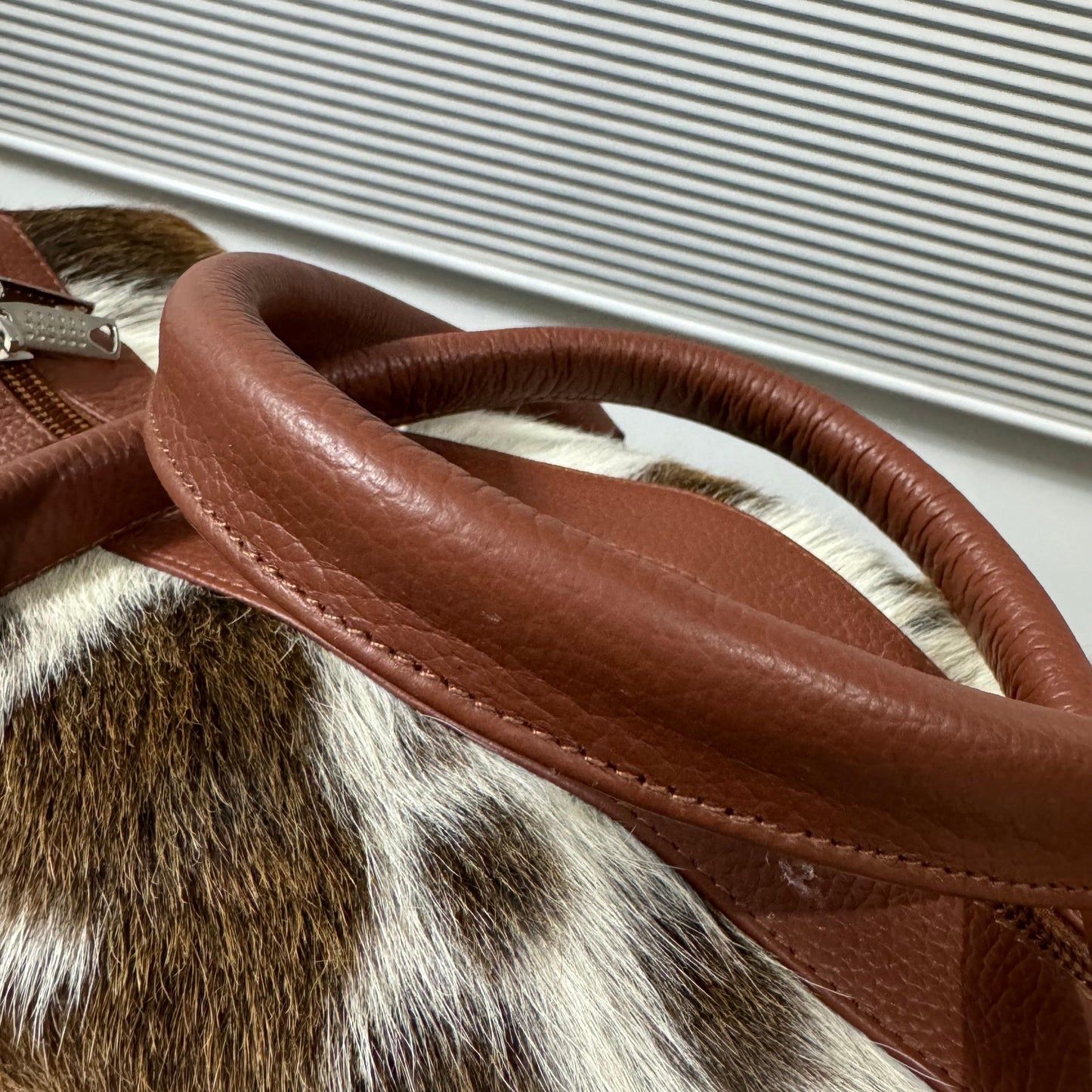 Close-up of a brown leather handbag handle with silver-tone hardware, resting on a brown, white, and black spotted animal-print hide.