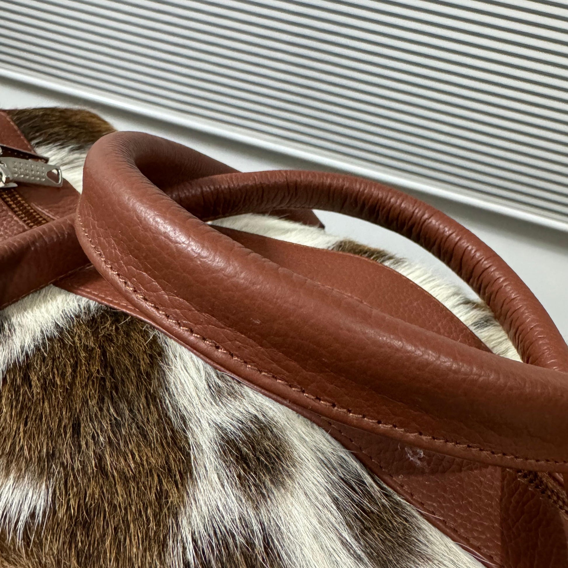 Close-up of a brown leather handbag handle with silver-tone hardware, resting on a brown, white, and black spotted animal-print hide.