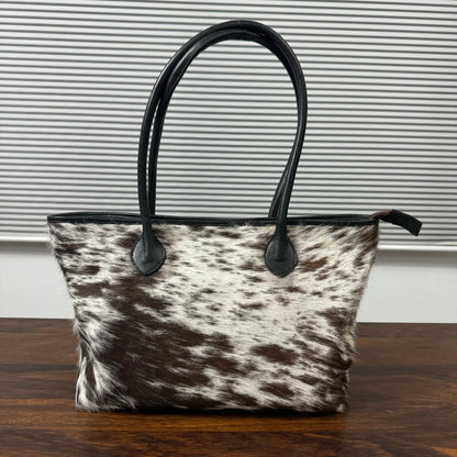 A cowhide tote bag with black leather handles and trim, sitting on a dark wooden table in front of a gray slatted window.