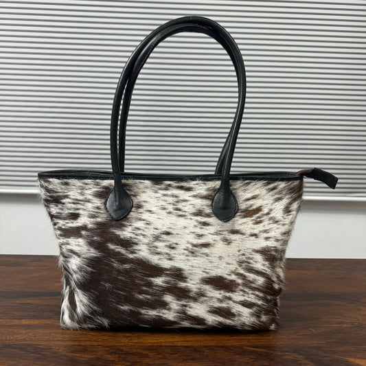 A cowhide tote bag with black leather handles and trim, sitting on a dark wooden table in front of a gray slatted window.
