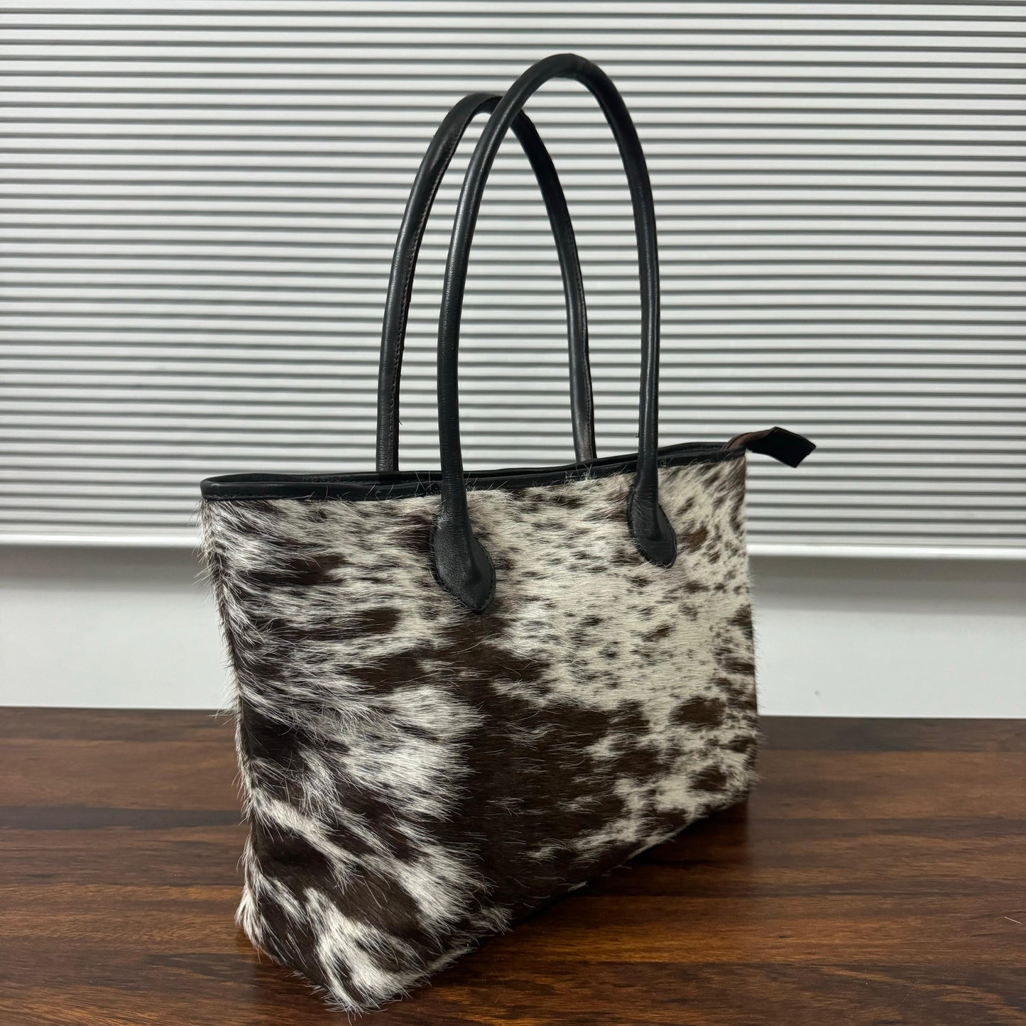 A cowhide tote bag with black leather handles, standing on a dark wooden table in front of a gray slatted window.