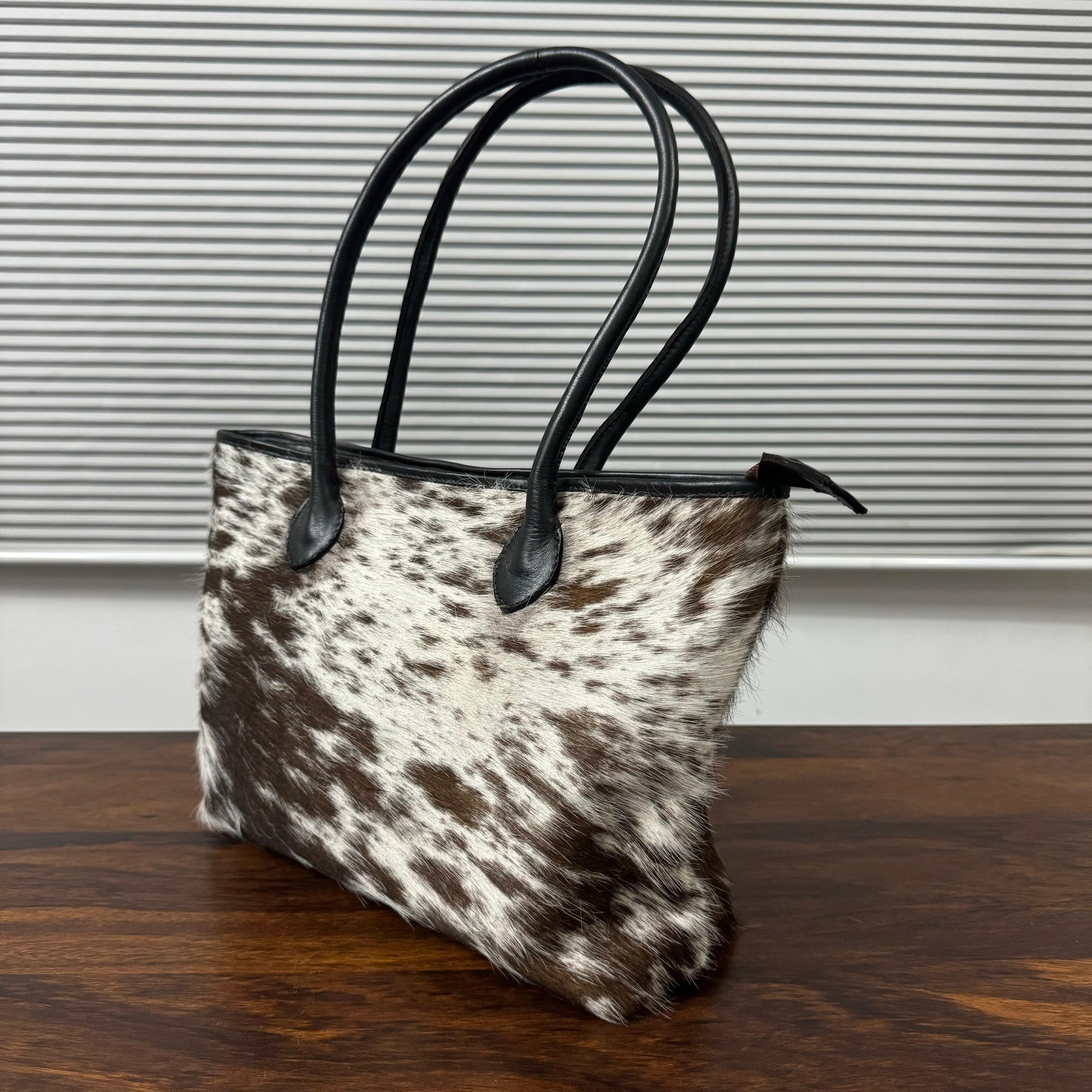 A cowhide handbag with a brown and white spotted pattern and black leather handles, resting on a wooden table in front of a gray slatted background.